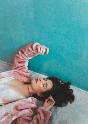 A girl with dark hair, wearing faux fur pink coat and pink top, lying down on a carpeted floor.