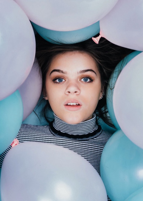 Upclose photo of a young girl with dark hair and blue eyes, whose face is surrounded by pink and blue balloons.