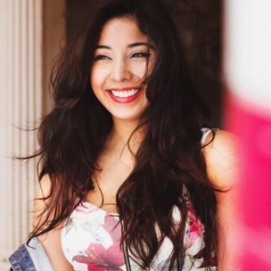 Shot of young girl, from the chest up, smiling while facing the camera. She is wearing a floral top with pink and red hues.