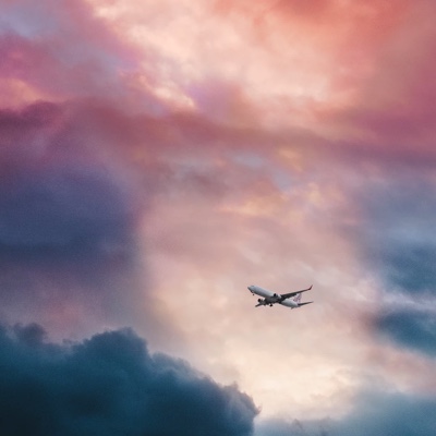 Image of a plane flying across a cloudy sky with some pink hues.